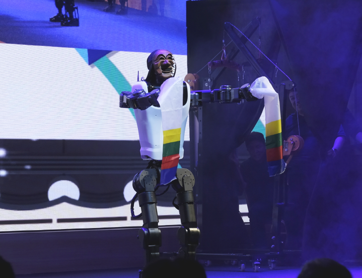 Humanoide KAIROS com máscara e tecidos coloridos nos braços durante apresentação em palco.